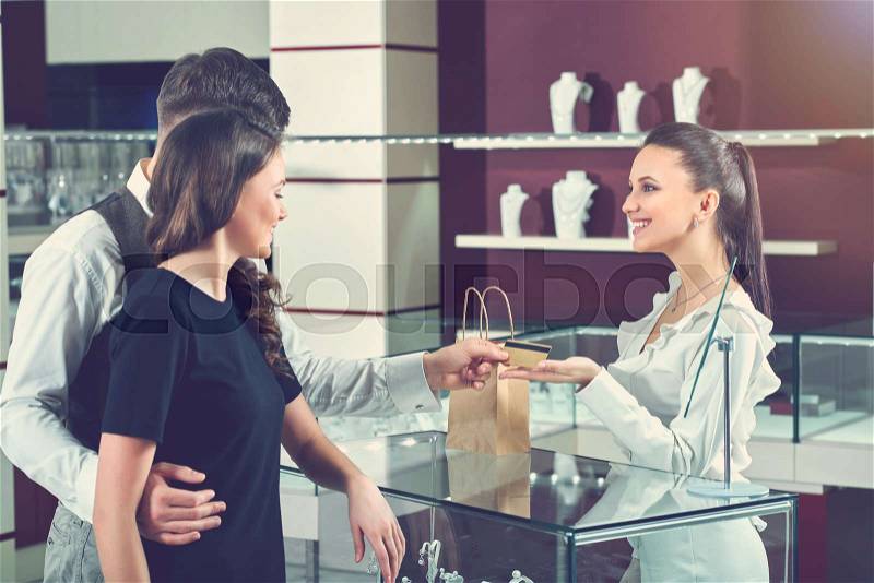 Couple of brunette girlfriend and boyfriend buying jewelry and paying for purchase. Jeweler seller happy looking, smiling to buyers and taking golden credit card for payment. Luxury jewelry store, stock photo