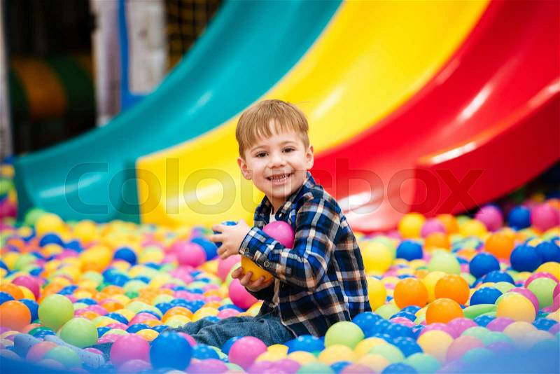 Cheerful little boy sitting and playing at indoor playground with colorful balls, stock photo