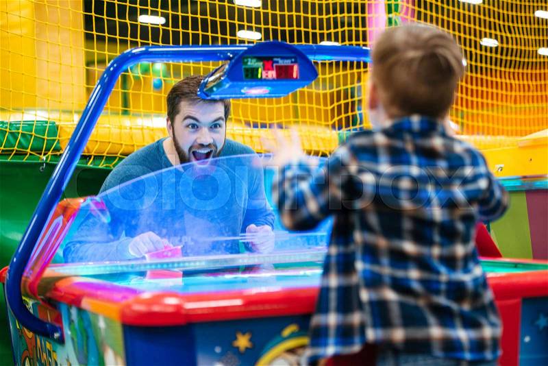 Joyful bearded father and son playing air hockey game at amusement park, stock photo