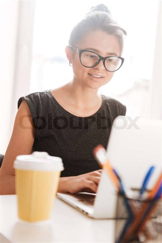 Concentrated charming young woman in glasses drinking coffee and working with laptop, stock photo