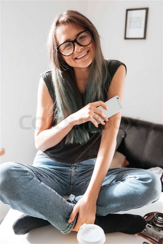 Smiling beautiful young woman sitting with legs crossed and using cell phone on the table, stock photo