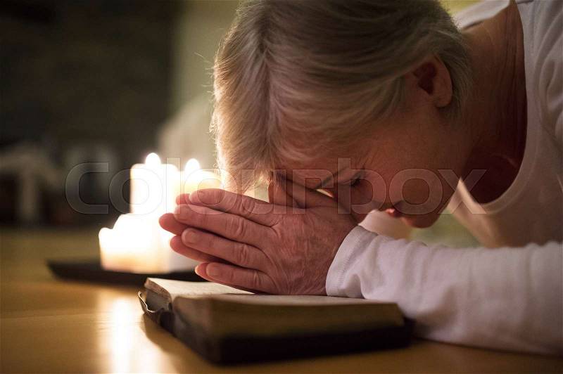 Senior woman kneeling on the floor praying with hands clasped together on her Bible. Burning candles next to her. Close up, stock photo