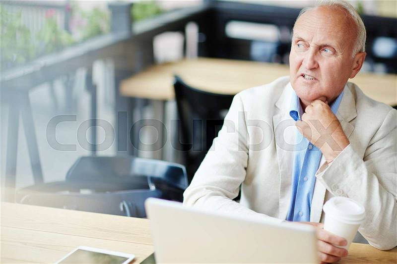 Frowning senior man looking sideways thinking hard while working with laptop at outdoor area of street cafe during coffee break, stock photo