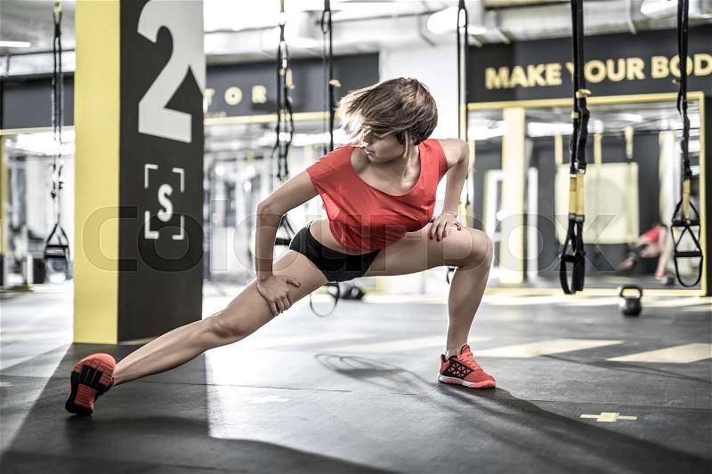 Delightful girl does stretching in the gym on the background of the hanging TRX straps. She wears a red sleeveless and sneakers, black shorts. Horizontal, stock photo