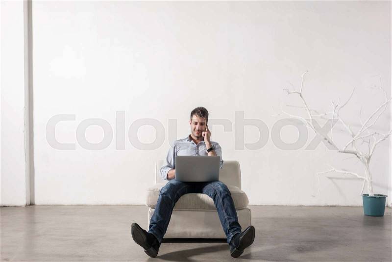 Young man sitting in empty room, using cell phone and notebook PC, stock photo