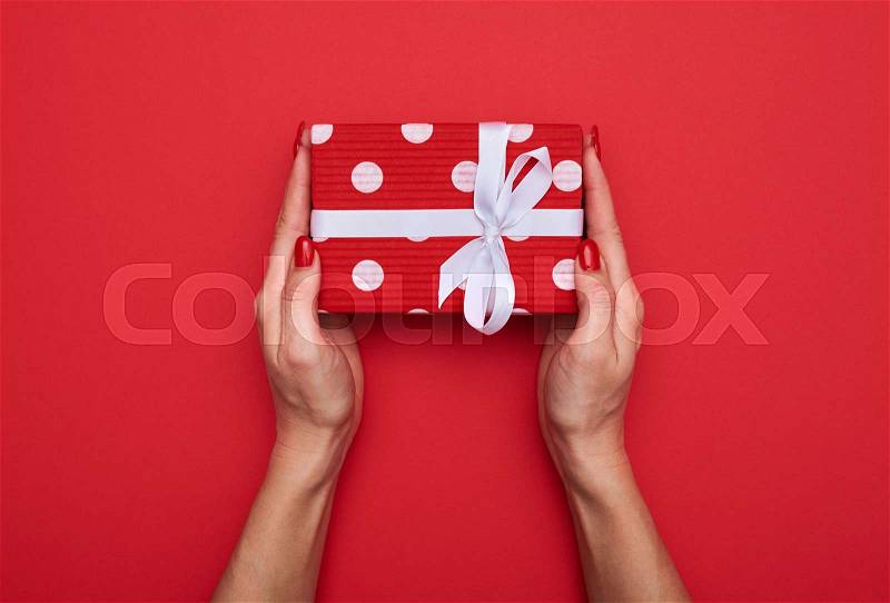 High angle of cute designed polka dot gift box with white ribbon in hands of female isolated over red background , stock photo