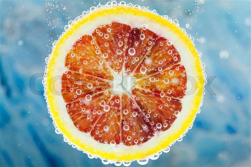 Close-up of slice of citrus blood orange dropped into carbonated water with bubbles on blue background, stock photo