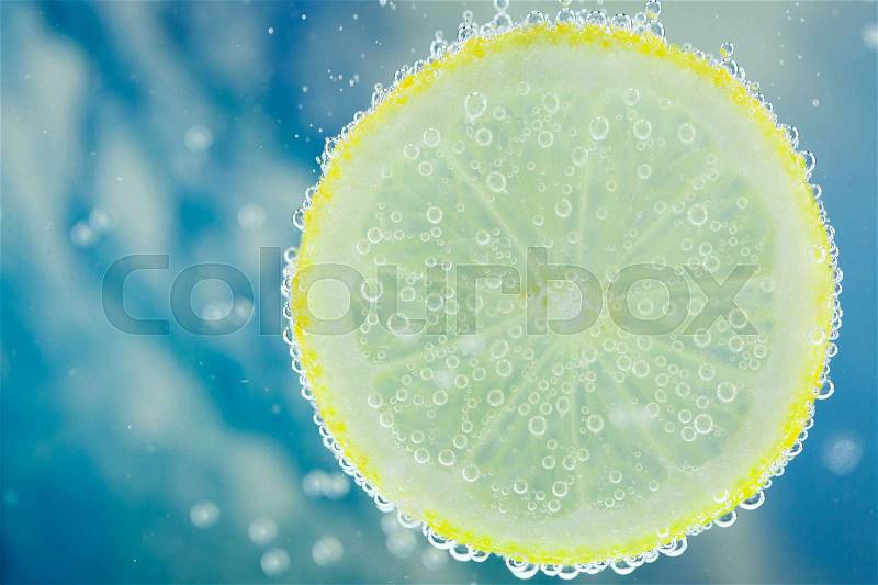 Vertical close-up of slice of lemon falling into carbonated water with bubbles, on blue background. Refresher drink concept, stock photo