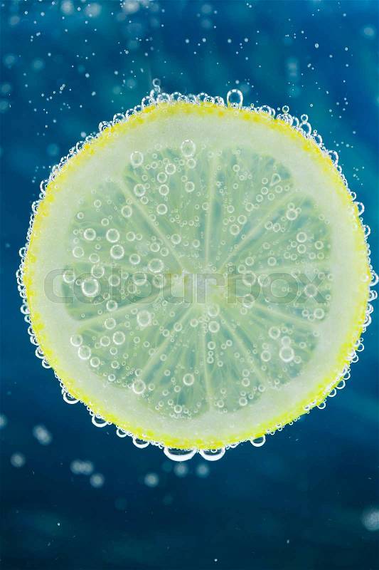 Vertical close-up of slice of lemon falling into carbonated water with bubbles, on blue background. Refresher drink concept, stock photo