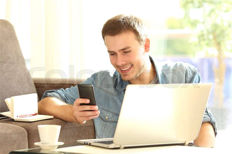Entrepreneur working on line with a smart phone and a laptop sitting on the floor of the living room at home, stock photo