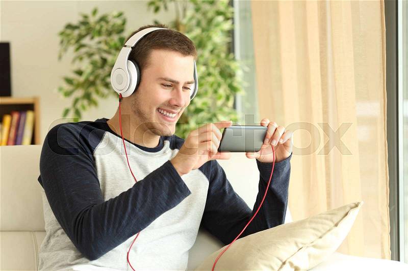 Handsome guy listening and watching media content on line in a smartphone sitting on a sofa in the living room at home, stock photo