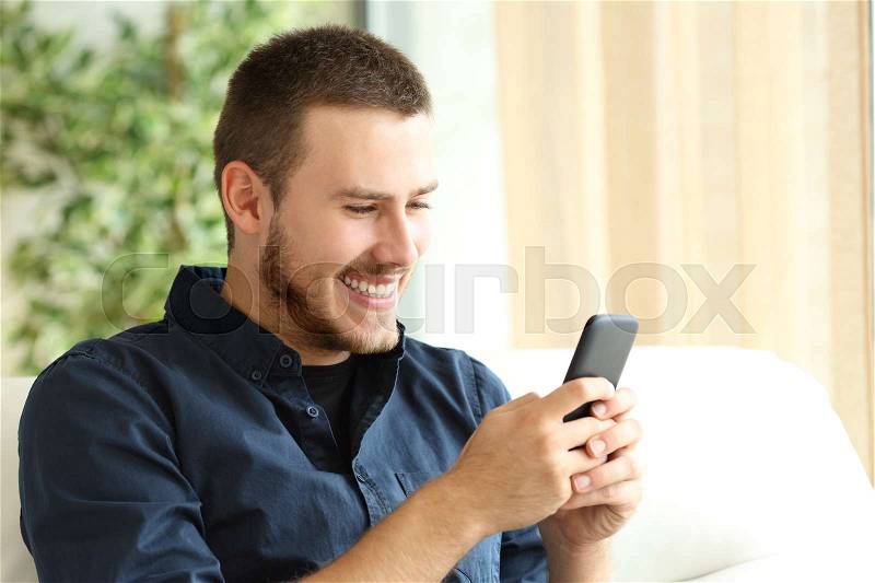 Portrait of a happy casual guy using a mobile phone sitting on a sofa in the living room at home with a window in the background, stock photo
