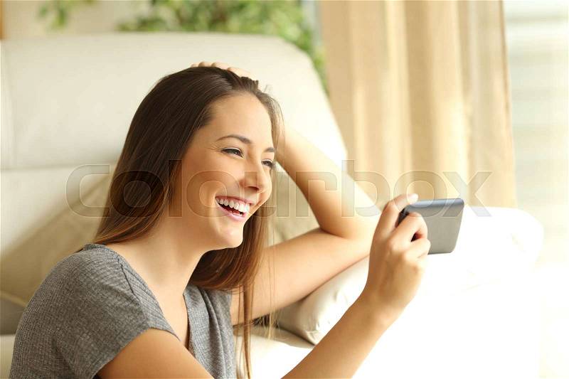 Girl watching streaming video in a smart phone sitting on the floor of the living room at home near a window, stock photo