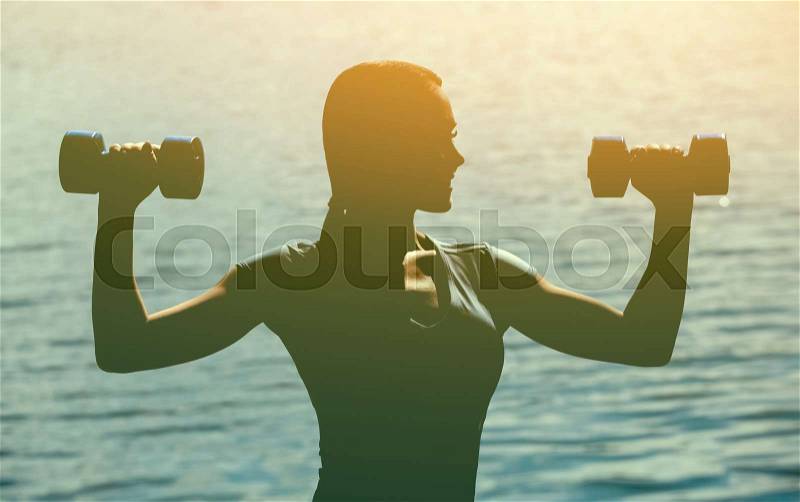 Silhouette of a beautiful athlete involved in sports, conducts training with dumbbells in hand, against the background of sunset at the lake. The concept of a healthy lifestyle and sports culture, stock photo