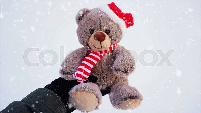 Female hand holding a cute Christmas teddy bear against snow on the ground with snowfall effect, stock photo