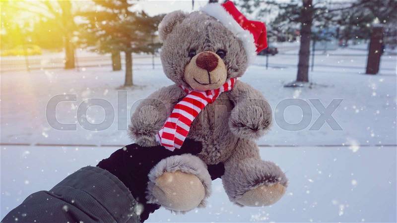 Female hand holding a cute Christmas teddy bear against winter background with sun ray and snow effect, stock photo