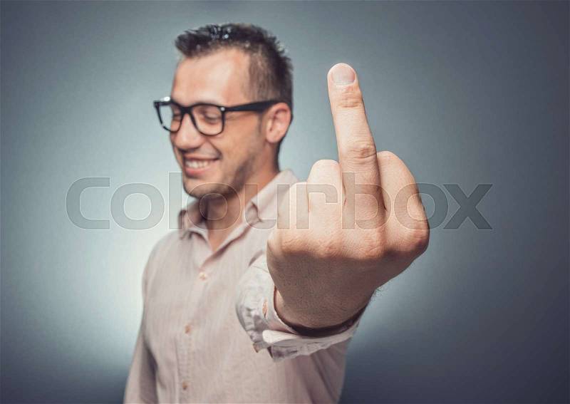 Smiling man giving the middle finger over gray dark background. Displeased young businessman, stock photo