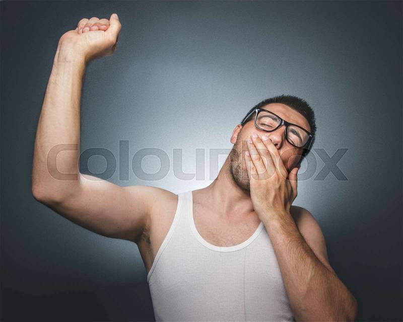 Man yawning. Awakening. Sleepy funny young guy wearing white undershirt over dark gray background, stock photo