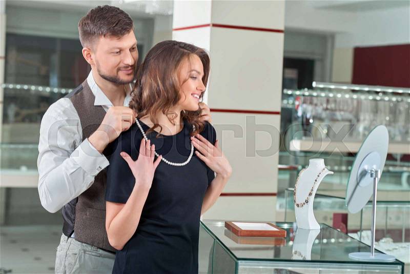 Couple of brunette woman with wavy hair wearing black dress and handsome man, trying necklace at jewelry store. White pearl necklace on neck of gorgeous girl. Looking at mirror and smiling each other, stock photo