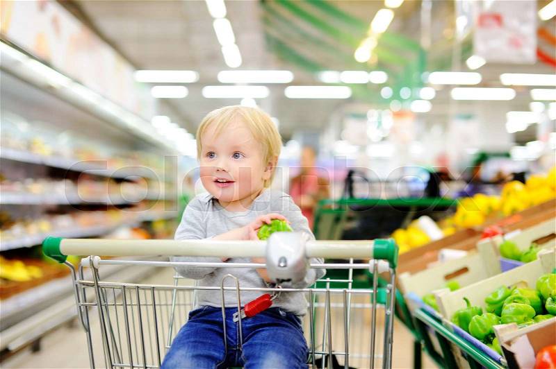 Cute toddler boy sitting in the shopping cart in a food store or a supermarket, stock photo