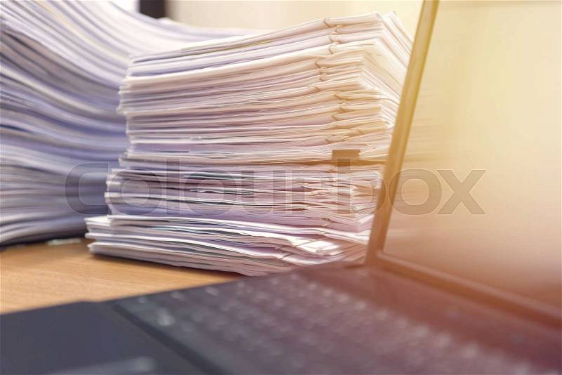 Computer notebook and documents on desk stack up high waiting to be managed, stock photo