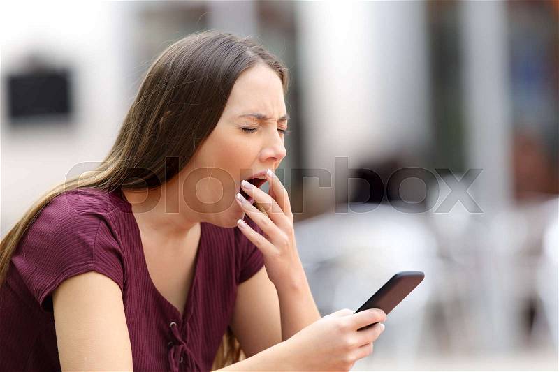 Woman yawning bored with on line content holding a mobile phone sitting on a bench in the street, stock photo