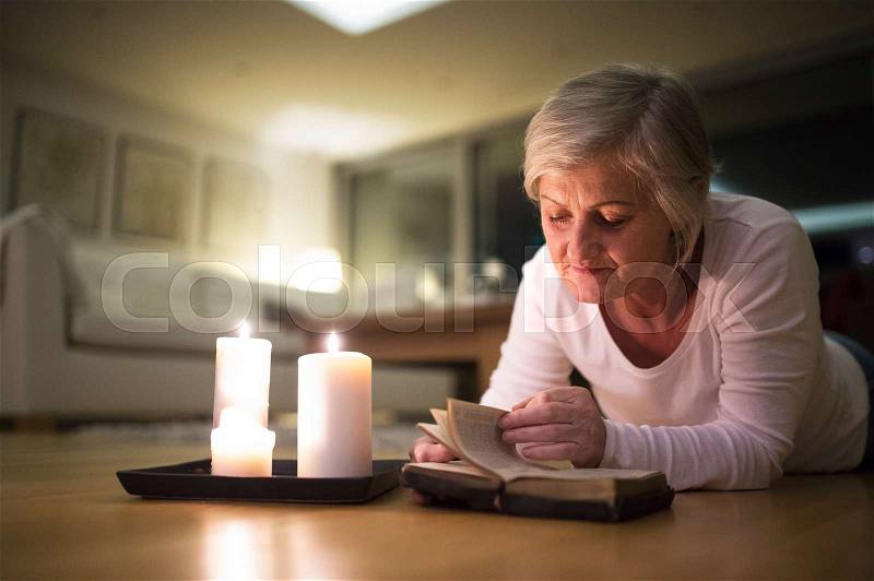 Beautiful senior woman at home in her living room lying on the floor reading her Bible. Burning candles next to her. Close up, stock photo