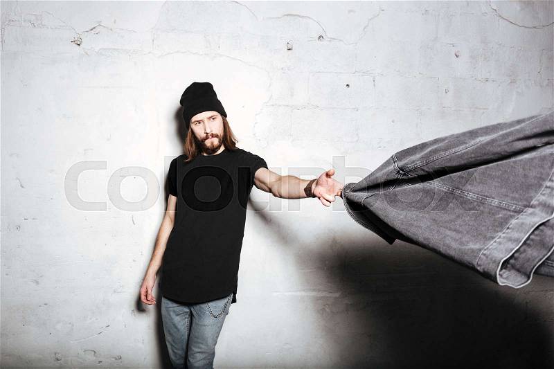Image of a young hipster man wearing hat standing isolated over wall background while holding jeans jacket, stock photo