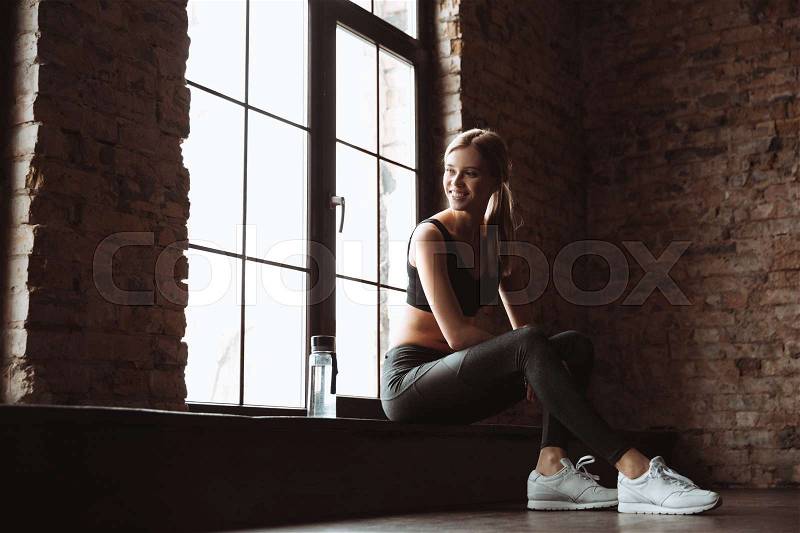 Photo of beautiful young fitness lady sitting in gym near bottle of water and window. Look aside, stock photo