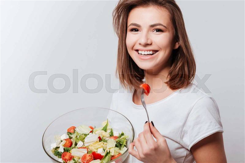 Photo of laughing young lady standing over white background while eating salad, stock photo
