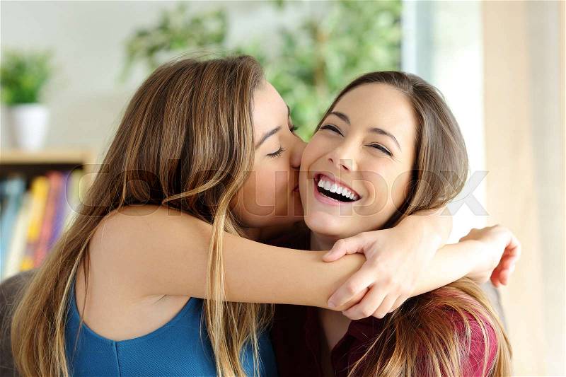 Affectionate girl kissing her happy sister or friend in the living room at home with a homey background, stock photo