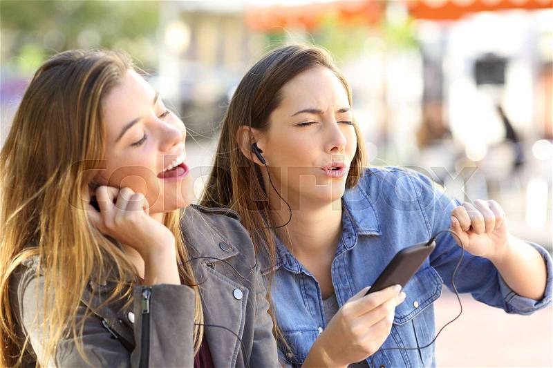 Joyful friends listening to music on line from a smart phone and singing sharing earphones in the street, stock photo