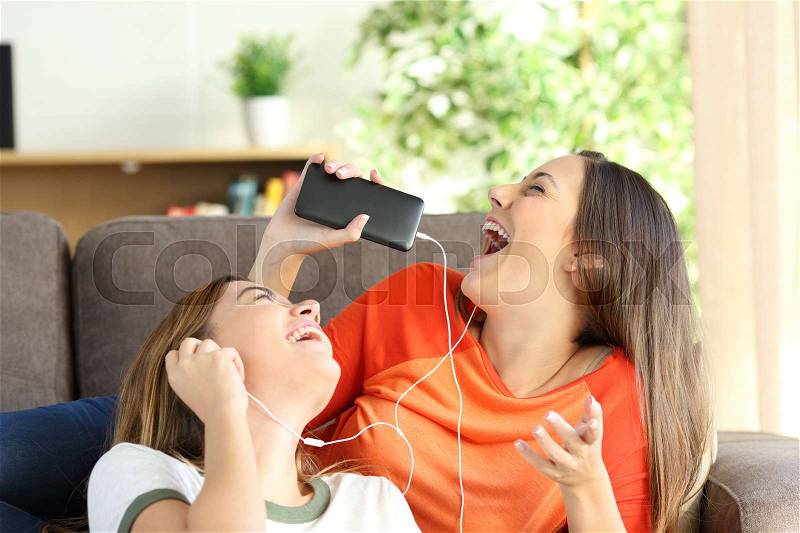 Excited friends or sisters listening music and singing on a couch in the living room at home, stock photo
