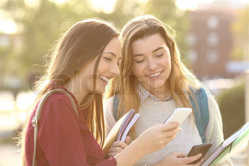 Two beautiful students watching media content on line in a smart phone outdoors in a park or university campus, stock photo