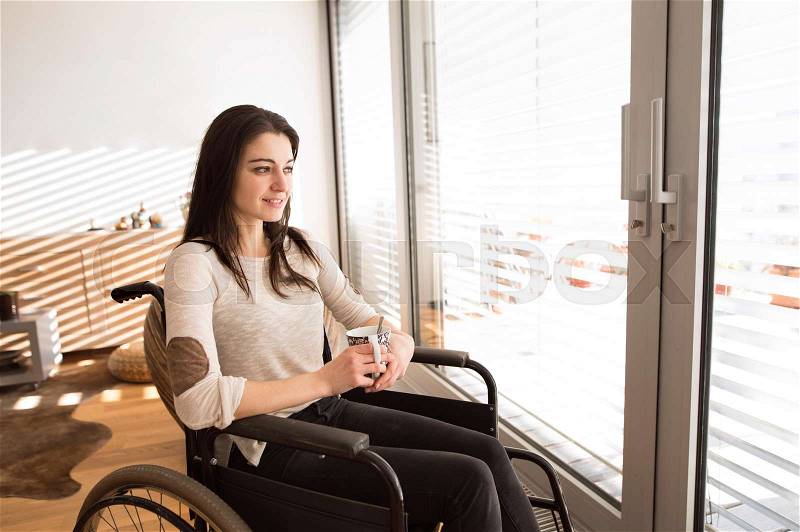 Beautiful young disabled woman in wheelchair at home in her living room, looking out the window, holding a cup of tea or coffee, stock photo