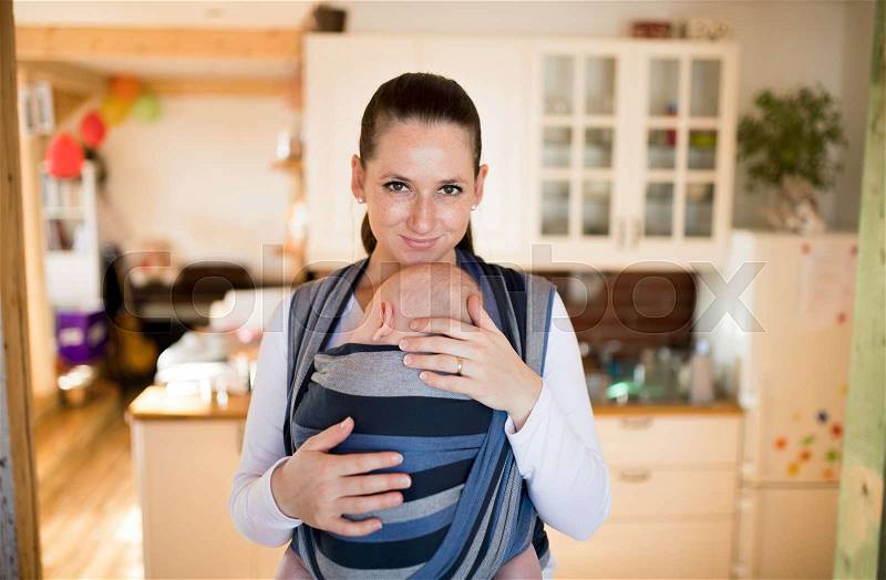 Beautiful young mother in kitchen with her baby son sleeping in sling at home, gently stroking him, stock photo