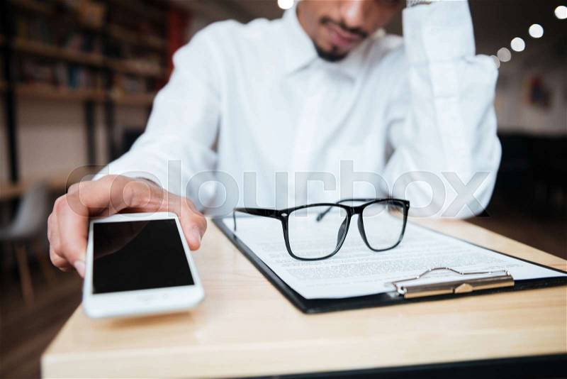 Cropped image of handsome african businessman sitting at the table holding phone. Coworking, stock photo