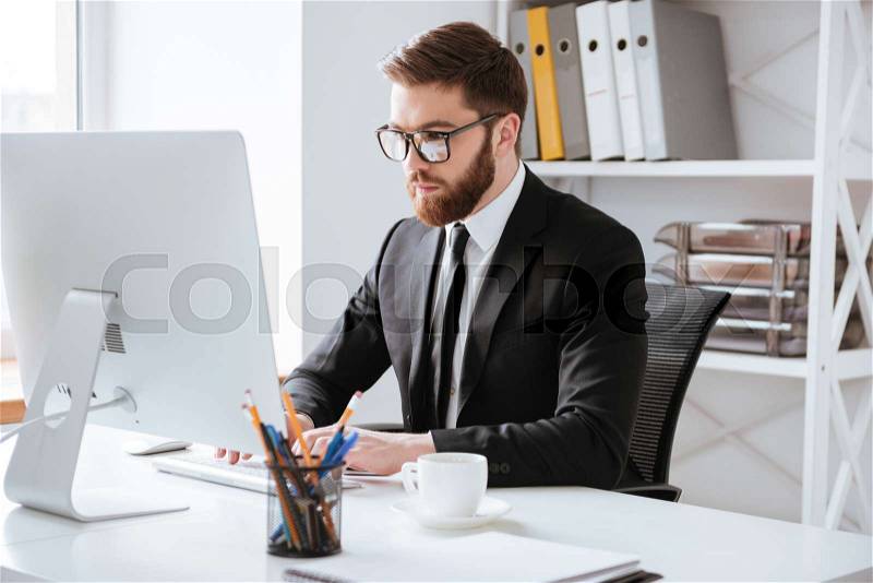 Image of attractive young bearded businessman sitting in office while using computer and looking aside, stock photo