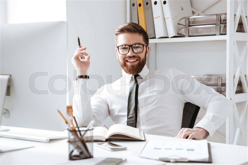 Photo of happy businessman sitting in office while writing notes. Looking at camera, stock photo