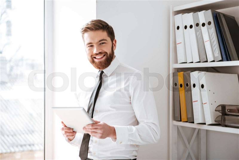 Image of happy young businessman dressed in white shirt standing in office holding tablet computer in hands and look at camera, stock photo