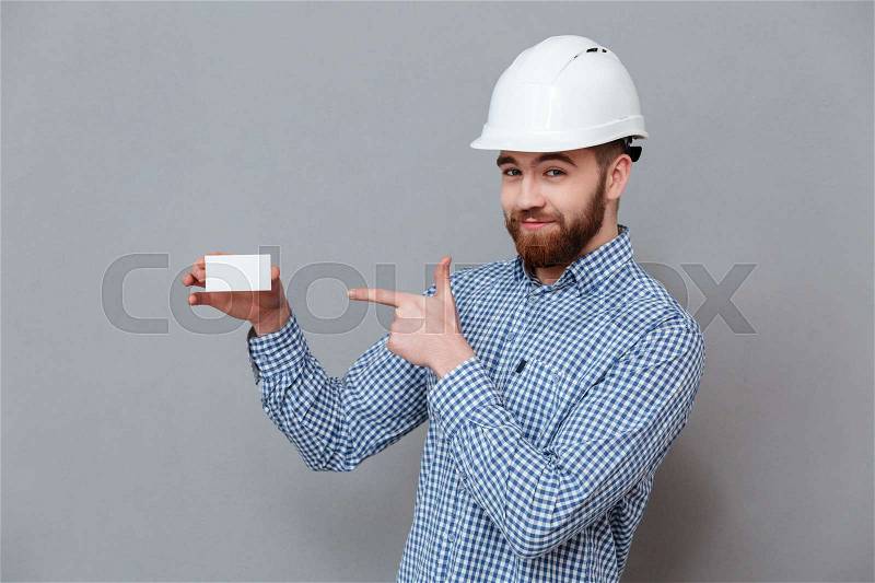Photo of cheerful bearded builder holding copyspace business card and pointing standing over grey background, stock photo