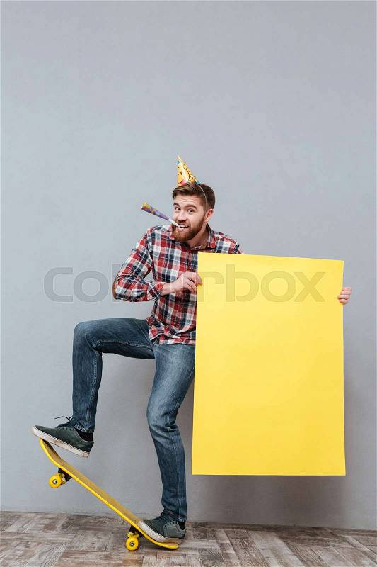 Photo of handsome young bearded birthday man on skateboard holding copyspace board standing over grey background, stock photo