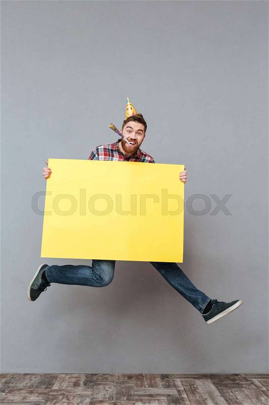 Picture of happy young bearded birthday man holding copyspace board jumping over grey background, stock photo