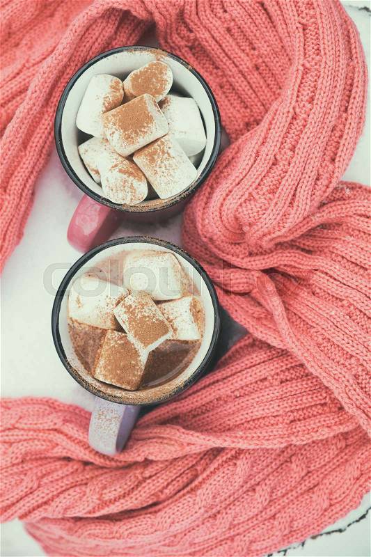 Hot chocolate with marshmallow in pink and violet two cups wrapped in a cozy winter pink scarf on the snow-covered table in the garden. Coloring and processing photo, small depth of field, stock photo