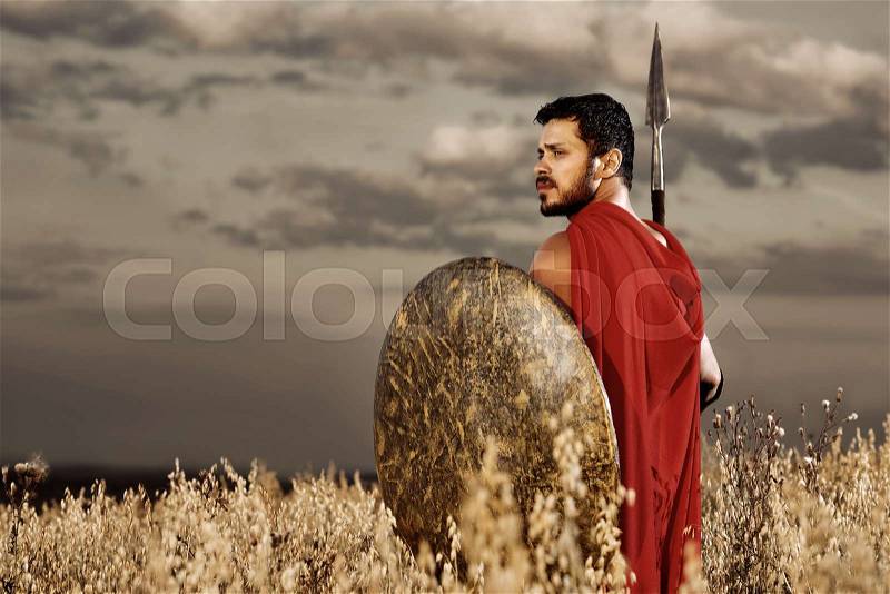 Back view of one warrior wearing like spartan confidently turned and looking away over shoulder, going in attack. Brunet with beard holding iron sword and shield. Bad weather with dark sky at field, stock photo