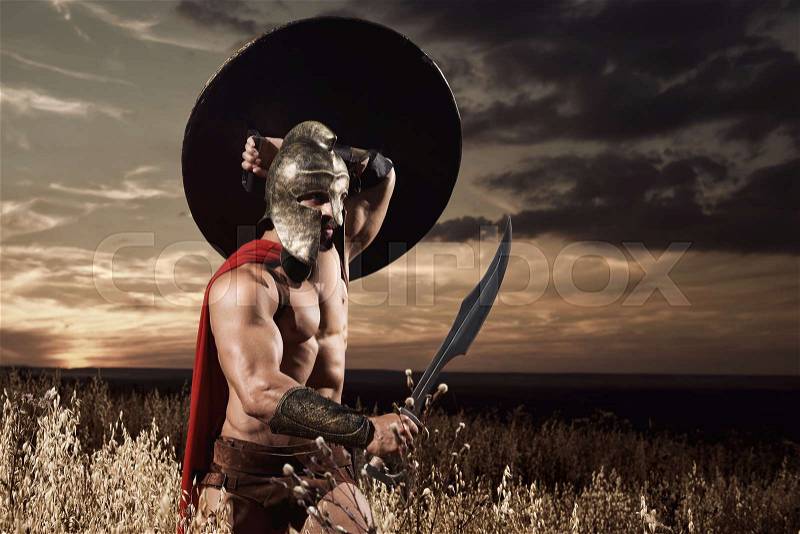 Warrior wearing in red cloak like spartan or antique roman soldier going forward in attack with iron sword. Incognito in helmet with bare torso holding bronze sword over head. Dark sky at sunset, stock photo