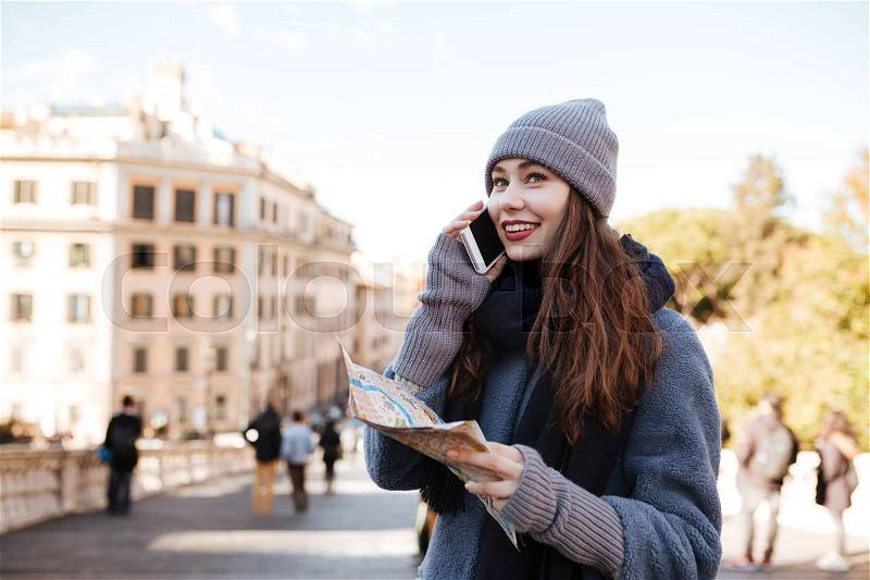 Cheerful beautiful young woman with map talking on mobile phone in the city, stock photo