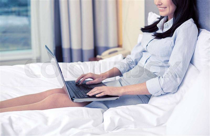 Business trip, people and technology concept - smiling businesswoman with laptop computer typing in bed at hotel, stock photo