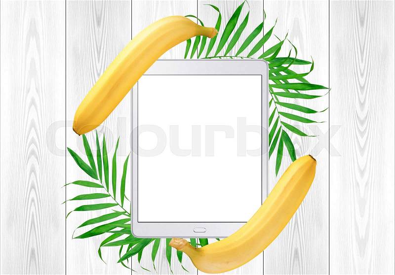 Tablet pc in green fern leaves and banana frame on white wood table. Top View, stock photo