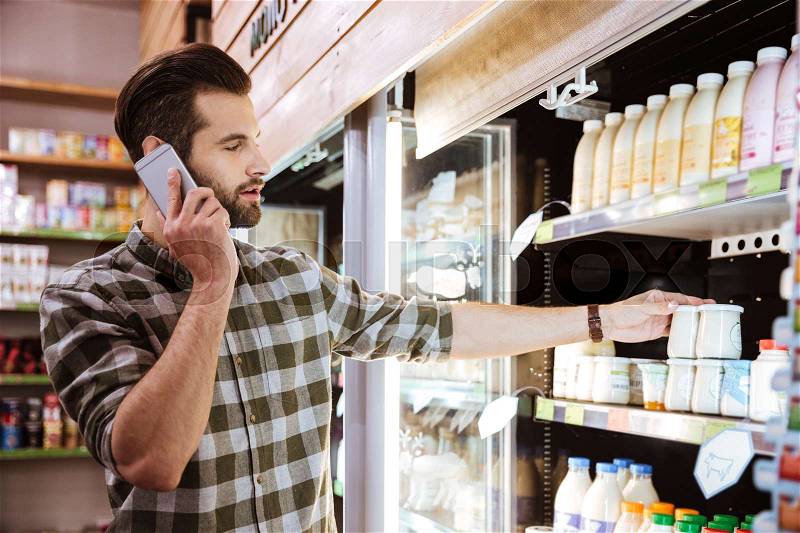 Handsome bearded young man talking on mobile phone at shopping in grocery shop, stock photo